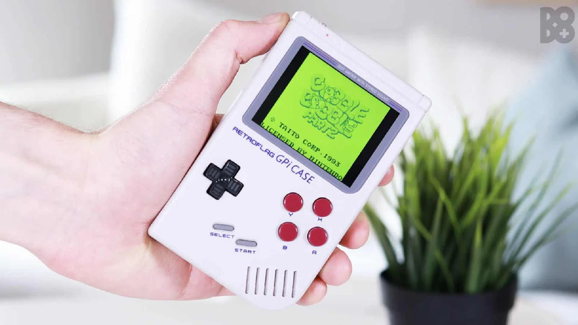 Console rétro portable façon Game Boy tenue en main, écran vert allumé, plante en arrière-plan
